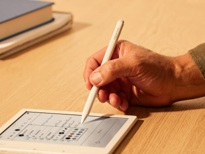 Une main tient un Kobo Stylet 2 au-dessus d'une liseuse Kobo Libra Colour posée sur une surface plane. La pointe du stylet est sur le point de sélectionner l'une des couleurs de stylo affichées dans un menu d'options sur l'écran de la liseuse.