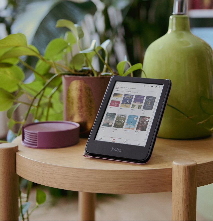 Une liseuse Kobo Clara Colour sur son support SleepCover, posée sur une table en bois avec une plante en pot et une lampe.