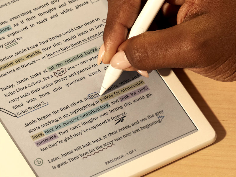 Une main utilise le Kobo Stylet 2 pour écrire directement dans un eBook sur une liseuse Kobo Libra Colour posée sur une surface plane. Sur la page, nous voyons des annotations et des surlignages colorés.