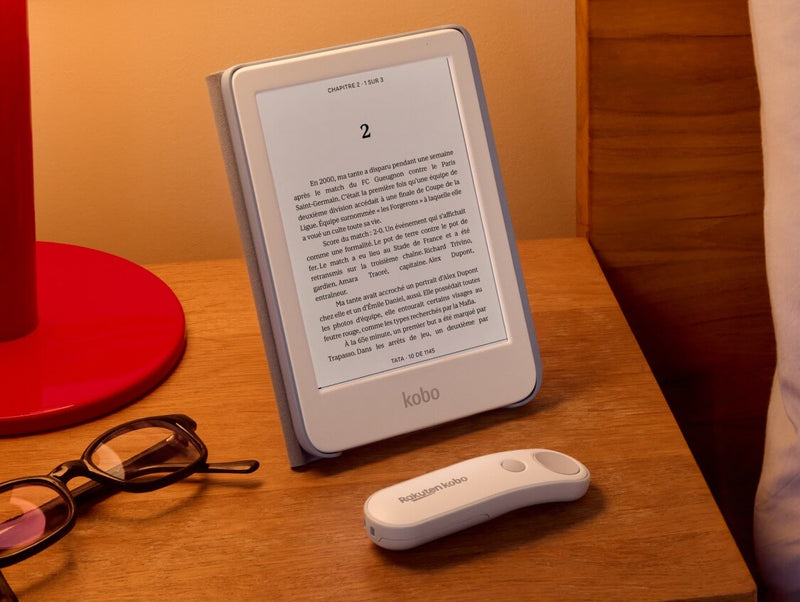 Une Kobo Clara Colour blanche est posée sur une table dans son étui SleepCover avec support intégré, à côté d'une Télécommande Kobo blanche et d'une paire de lunettes.