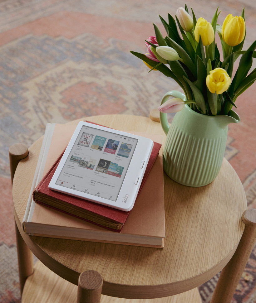 Une liseuse Kobo Libra Colour sur deux livres reliés, posés sur une table en bois à côté d’un vase vert contenant des fleurs.